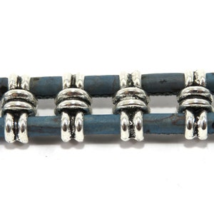 K&ouml;nnte beinhalten: Vier silberne Metallperlen mit einem dekorativen Knotendesign sind auf einer doppelten Schnur aus blauem Kork gef&auml;delt.