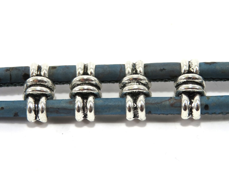 K&ouml;nnte beinhalten: Vier silberne Metallperlen mit einem dekorativen Knotendesign sind auf einer doppelten Schnur aus blauem Kork gef&auml;delt.