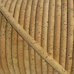 Puede incluir: Primer plano de un rollo de cinta de corcho natural con un borde cosido decorativo. El corcho es de color marrón claro con manchas más oscuras, y el borde es de color beige claro. La cinta se utiliza probablemente para manualidades o decoración.