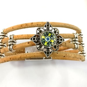 K&ouml;nnte beinhalten: Ein mehrreihiges Armband aus Kork mit einem silberfarbenen, dekorativen Verschluss, der mit einem blauen, gelben und gr&uuml;nen gemusterten Glascabochon verziert ist.