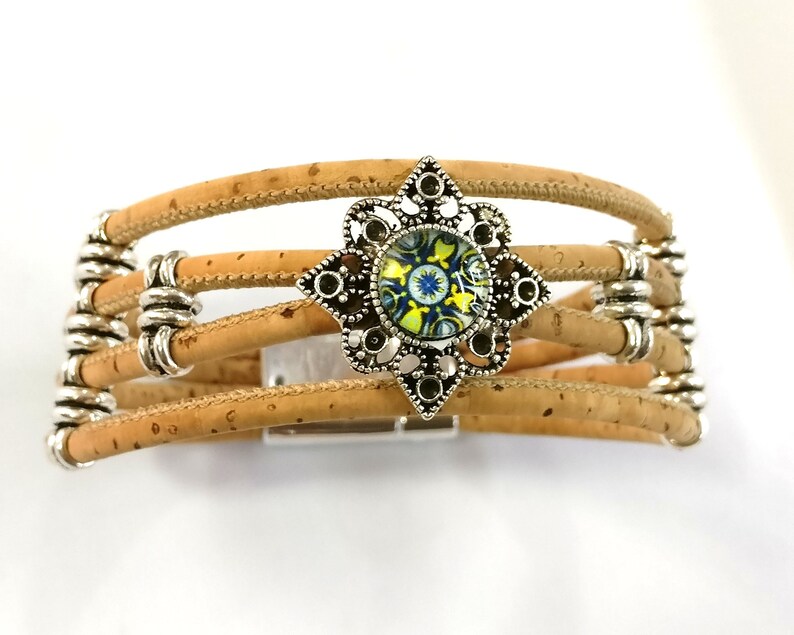K&ouml;nnte beinhalten: Ein mehrreihiges Armband aus Kork mit einem silberfarbenen, dekorativen Verschluss, der mit einem blauen, gelben und gr&uuml;nen gemusterten Glascabochon verziert ist.