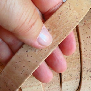 May include: A close-up of a stack of cork strips, each with a unique pattern of brown and beige flecks. The strips are held together by a hand, showcasing the natural texture of the cork.