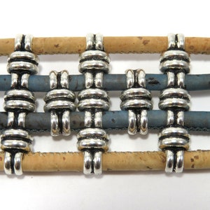 K&ouml;nnte beinhalten: Silberne Metall-Kordelenden f&uuml;r Armb&auml;nder oder Halsketten. Die Kordelenden sind so konzipiert, dass sie mehrere Str&auml;nge aus Kordel, Leder oder Stoff halten. Die Kordelenden sind auf einem Hintergrund aus Kork und blauem Leder abgebildet.