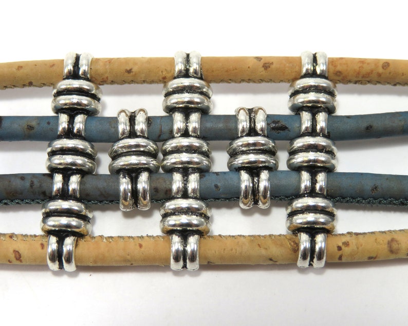 K&ouml;nnte beinhalten: Silberne Metall-Kordelenden f&uuml;r Armb&auml;nder oder Halsketten. Die Kordelenden sind so konzipiert, dass sie mehrere Str&auml;nge aus Kordel, Leder oder Stoff halten. Die Kordelenden sind auf einem Hintergrund aus Kork und blauem Leder abgebildet.