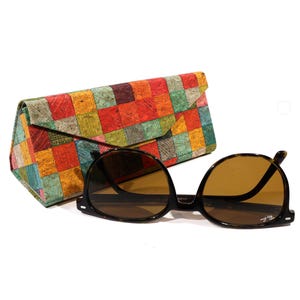 May include: A pair of round sunglasses with brown lenses and black frames, alongside a colourful, patchwork-style glasses case. The case has a geometric pattern in red, orange, green, and blue. The sunglasses have a classic, retro design.