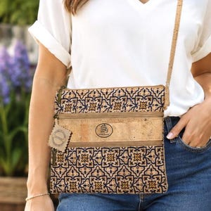 Puede incluir: Un bolso bandolera rectangular de corcho con un patrón geométrico repetitivo en azul y dorado. El bolso cuenta con dos compartimentos con cremallera, una correa fina de corcho y un pequeño parche circular con el logo de un barco.