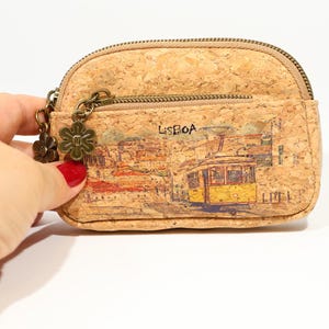 May include: Cork coin purse with a zipper closure and a small front pocket. The purse features a printed design of a yellow tram in Lisbon, Portugal, with the word "LISBOA" above it. A flower charm hangs from the zipper.