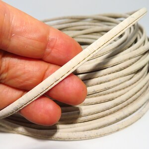 Puede incluir: Primer plano de un cordón de corcho blanco con un hilo dorado que recorre el borde. El cordón está enrollado y se sostiene en una mano.