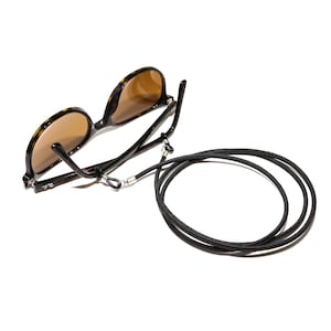 Pode incluir: Óculos de sol com padrão de tartaruga, lentes castanhas e uma corrente de cordão preto. A corrente possui conectores de metal prateado que se prendem às extremidades das hastes dos óculos. O logótipo Ray-Ban é visível na haste.