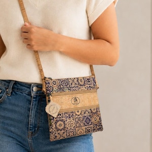 May include: A rectangular crossbody bag made of natural cork material featuring a patterned design of blue, white, and tan tiles. The bag includes a front zippered pocket, a thin adjustable cork strap, and a small circular logo on the front panel.
