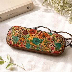 May include: A rectangular eyeglass case with a cork texture surface featuring a colorful floral print with red, teal, and yellow flowers, green leaves, and small insects. A pair of dark brown tortoiseshell glasses rests partially inside the case.