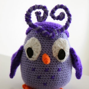 Puede incluir: Un búho de crochet morado con ojos blancos y un pico naranja. El búho tiene dos antenas de hilo rizado morado en la parte superior de su cabeza.