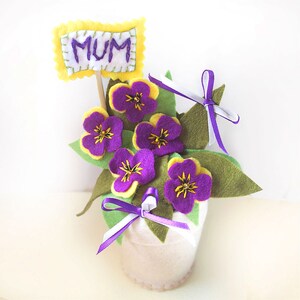 May include: Felt flower arrangement in a white pot with a yellow sign that says "MUM" and purple and yellow flowers with green leaves and purple ribbons.