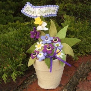 May include: A felt flower arrangement in a cream-colored pot with a purple ribbon. The flowers are purple, white, and yellow with green leaves. A banner above the flowers reads "Great Grandma".
