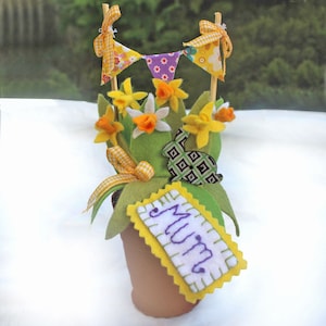 May include: A handmade felt flower arrangement in a terracotta pot. The arrangement includes yellow daffodils, green felt leaves, and a small bunny. A banner with yellow and purple flags sits above the flowers. A tag with the word "MUM" is attached.