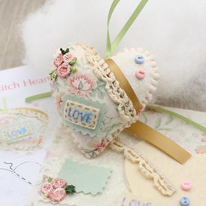 May include: A heart-shaped craft project with floral fabric, lace trim, and a gold ribbon. The word "LOVE" is embroidered on a small patch. Pink and blue buttons and a green ribbon are also visible. A fluffy white cloud-like material is in the background.