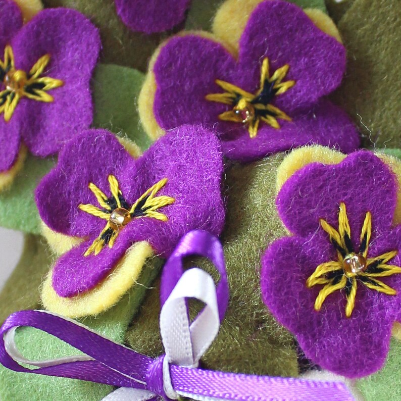 May include: A purple felt flower bouquet with yellow centers and embroidered details. The flowers are arranged in a circle and tied with a purple ribbon.