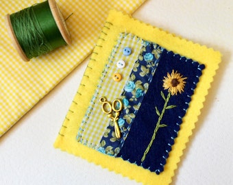 May include: A yellow felt pincushion with a blue and white floral fabric, a yellow and white gingham fabric, and a blue felt fabric with an embroidered sunflower. A pair of gold scissors are pinned to the fabric.