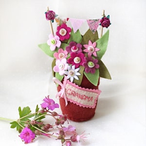 May include: A felt flower arrangement in a red pot with a pink banner that says "Grandma". The flowers are pink, white, and green.