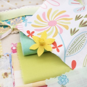 Puede incluir: Una colección de recortes de tela en varios colores y patrones, incluyendo estampados florales, diseños geométricos y colores sólidos. Una flor de fieltro amarilla se coloca encima de la tela.