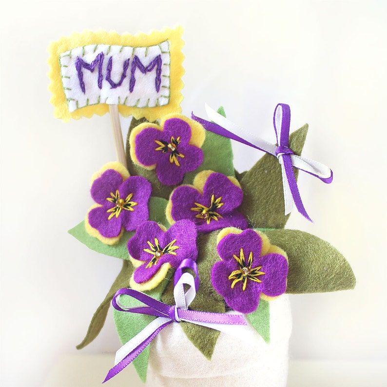 May include: A felt flower arrangement in a white pot with a yellow felt sign that says "MUM". The flowers are purple with yellow centers and green leaves.