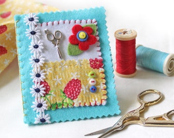 May include: A turquoise felt pincushion with a floral fabric patch, a pair of silver scissors, a red felt flower with yellow button center, and white daisy trim.