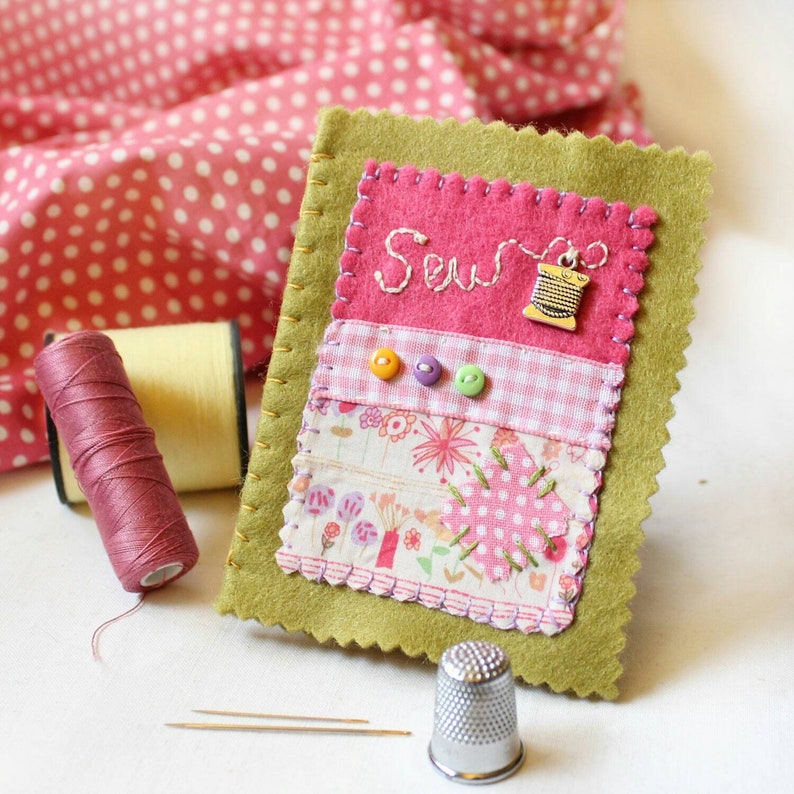 Handmade sewing needle book of green felt with pink applique design including the embroidered word 'sew', a silver cotton reel charm and three tiny buttons