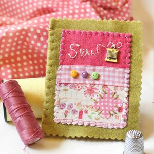 Handmade sewing needle book of green felt with pink applique design including the embroidered word 'sew', a silver cotton reel charm and three tiny buttons