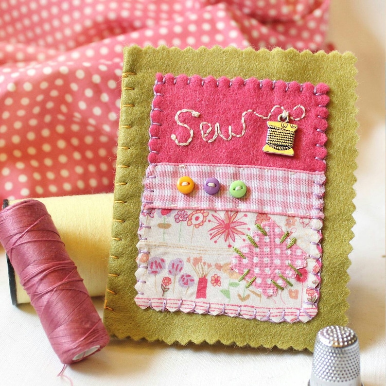 Handmade sewing needle book of green felt with pink applique design including the embroidered word 'sew', a silver cotton reel charm and three tiny buttons