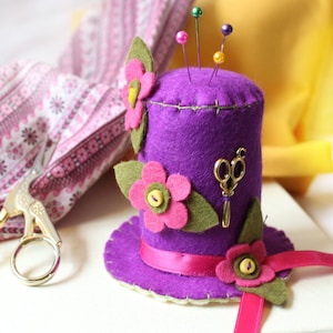 May include: Purple felt pincushion shaped like a hat with pink felt flowers, green leaves, and a gold pair of scissors. The hat has a pink ribbon around the base.