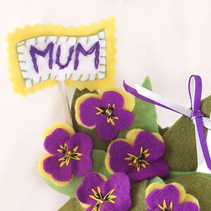 May include: A felt flower arrangement with purple and yellow flowers and green leaves. A yellow felt sign with the word "MUM" in purple embroidery is attached to a stick.