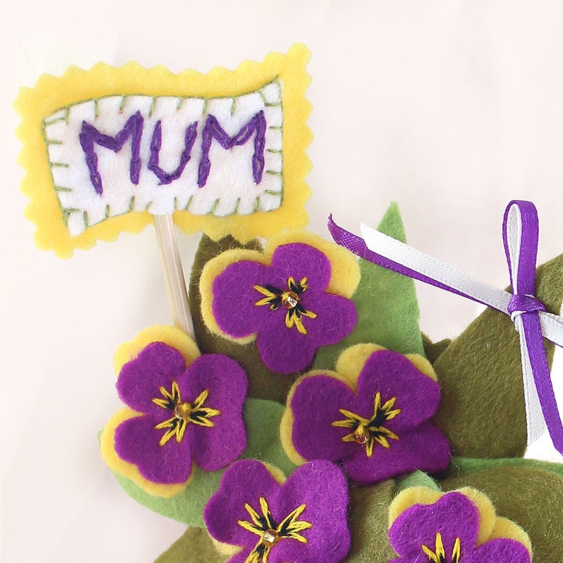 May include: A felt flower arrangement with purple and yellow flowers and green leaves. A yellow felt sign with the word "MUM" in purple embroidery is attached to a stick.