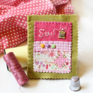 Handmade sewing needle book of green felt with pink applique design including the embroidered word 'sew', a silver cotton reel charm and three tiny buttons