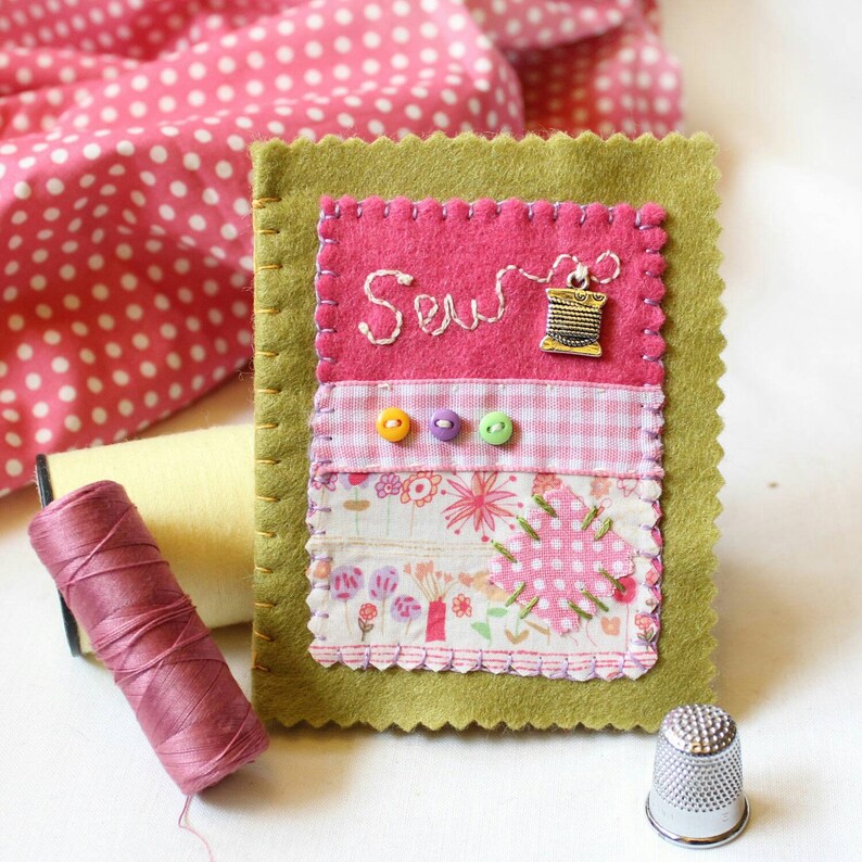 Handmade sewing needle book of green felt with pink applique design including the embroidered word 'sew', a silver cotton reel charm and three tiny buttons