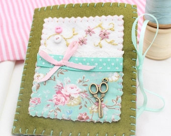 Puede incluir: Un alfiletero de fieltro verde con un diseño de tela floral, una cinta rosa y unas tijeras pequeñas. El alfiletero tiene un lazo de cinta azul claro para colgar.