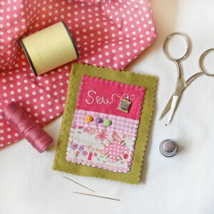 Handmade sewing needle book of green felt with pink applique design including the embroidered word 'sew', a silver cotton reel charm and three tiny buttons