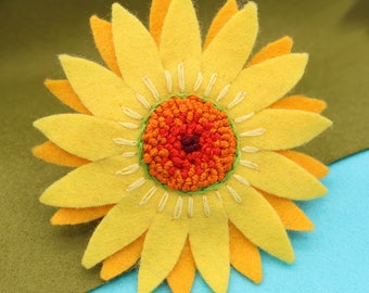 Könnte beinhalten: Eine Filz-Sonnenblumenbrosche mit einem gelben, orangefarbenen und roten Zentrum. Die Sonnenblume ist aus Filz gefertigt und hat einen mehrschichtigen Look.