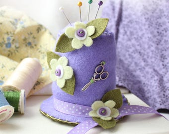 May include: Purple felt pincushion shaped like a hat with green felt leaves and white felt flowers. The hat has a purple and white polka dot ribbon around the brim. There are small silver scissors on the side of the hat.