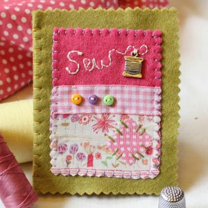 Handmade sewing needle book of green felt with pink applique design including the embroidered word 'sew', a silver cotton reel charm and three tiny buttons