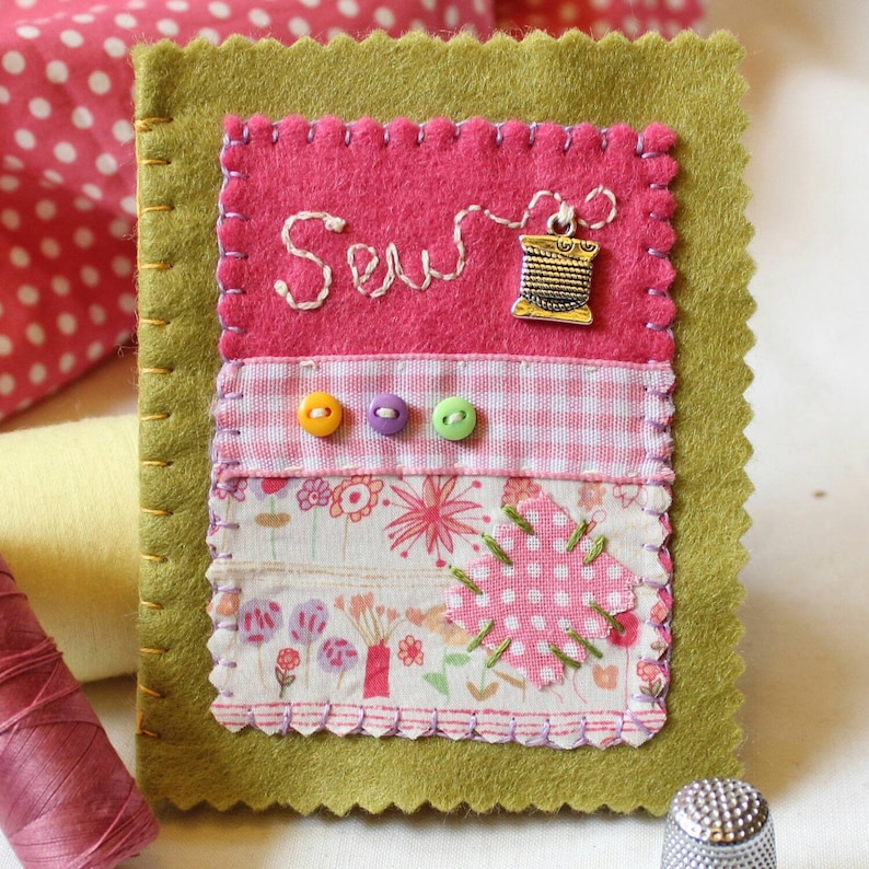 Handmade sewing needle book of green felt with pink applique design including the embroidered word 'sew', a silver cotton reel charm and three tiny buttons