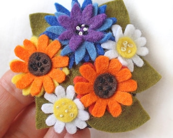 Könnte beinhalten: Eine Brosche in Form eines Blumenstraußes aus Filz mit orangefarbenen, blauen, gelben und weißen Blumen. Die Blumen sind auf einem grünen Filzgrund angeordnet.