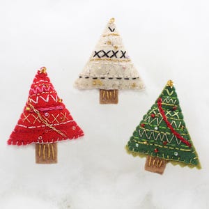 Könnte beinhalten: Drei handgefertigte Weihnachtsbaumornamente. Eines ist rot mit weißen und goldenen Nähten, ein anderes ist weiß mit schwarzen und goldenen Nähten, und das dritte ist grün mit roten und goldenen Akzenten. Jeder Baum hat einen braunen Stamm und eine goldene Perle oben.