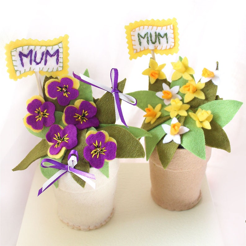 May include: Two felt flower arrangements in pots, each with a yellow felt flag that says "MUM". One arrangement has purple and yellow flowers, the other has yellow flowers.
