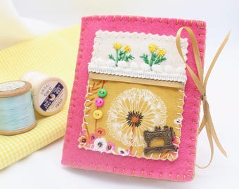 Puede incluir: Un libro de agujas de fieltro rosa con un bolsillo de tela amarilla que presenta un diseño de diente de león blanco y un pequeño amuleto de máquina de coser de metal. El bolsillo tiene un borde de encaje blanco y botones coloridos. Una cinta dorada está unida al libro.