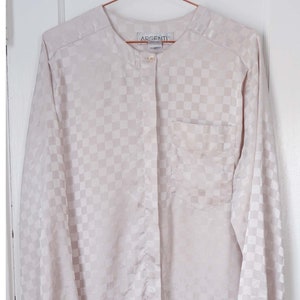 May include: A cream-colored, long-sleeved button-down shirt with a checkered pattern. The shirt has a mandarin collar and a single button closure at the neck. The shirt also has a patch pocket on the left chest. The brand label reads "Argenti".