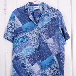 May include: A blue and white patterned short-sleeved button-up shirt with a collar. The shirt has a patchwork design with different floral and geometric patterns. The label on the shirt reads "Alfred Dunner".