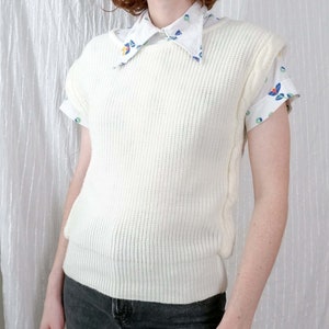 May include: A white knitted sleeveless sweater vest with a ribbed texture. The vest has a round neckline and is worn over a white collared shirt with a floral print.