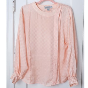 May include: A pale pink, long-sleeved blouse with a checkered pattern. The blouse has a round neckline and a button closure at the neck. The brand label reads "ARCENTI".
