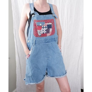 May include: Light blue denim overalls with a patchwork pocket on the front. The pocket is made of red, white, and blue fabric with a floral pattern.