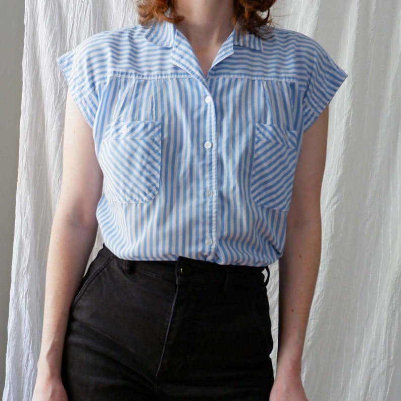 May include: A blue and white striped short-sleeved button-up shirt with a collared neckline. The shirt has two chest pockets and is tucked into black pants.